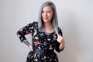 a picture of a woman, aleissia laidacker, standing with her hair dyed a sort of cotton candy grey and wearing a dress  with tattoo illustrations on it