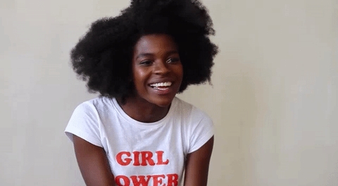 A smiling woman, African American, bouncing excitedly. She's wearing a white shirt with red text that reads "girl power"
